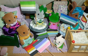 A group of Sylvanian Families animal figurines in a dollhouse bedroom, either holding aro-spec pride flags or sitting around a pile of pride flags on the floor. Flags include aro, fray, akoi/lith, cupio, demi and quoi, and animal figurines include two brown beavers, a grey rabbit, a green frog, a brown rabbit and a black-and-white cat baby. The grey rabbit, holding an aro pride flag, is wearing a striped dress in aro-pride colours.
